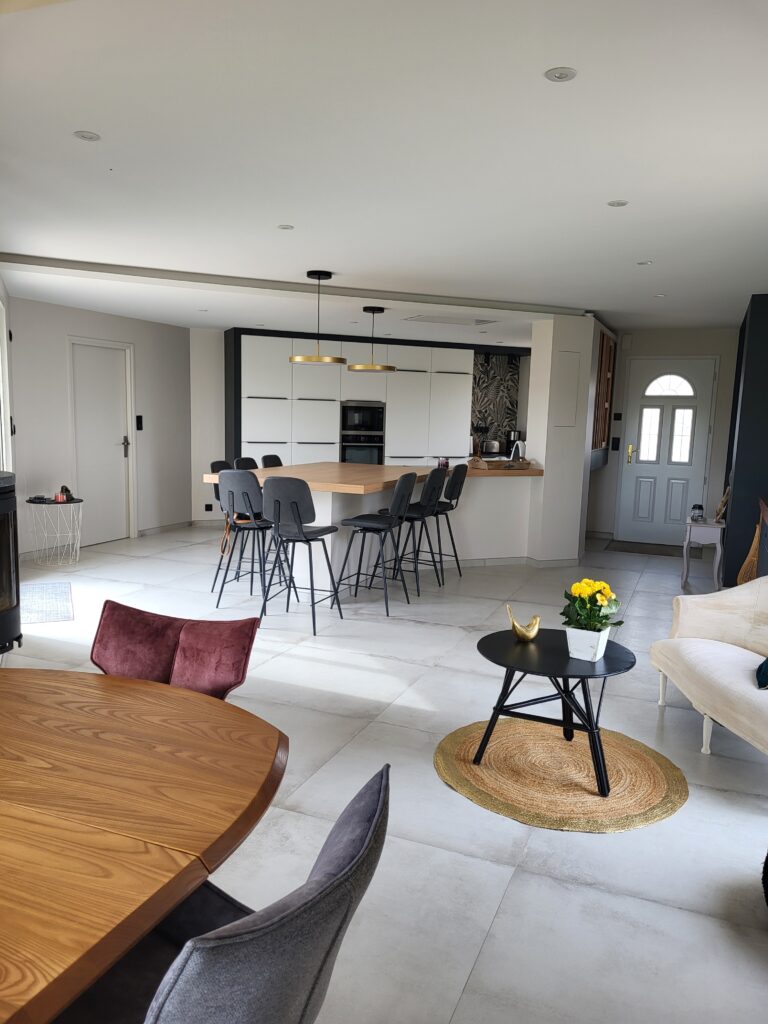 Cuisine et salle à manger modernes et ouvertes, avec un grand îlot, des chaises hautes, une table basse ronde avec des fleurs, des meubles aux tons neutres et un élégant carrelage pour une touche de style.