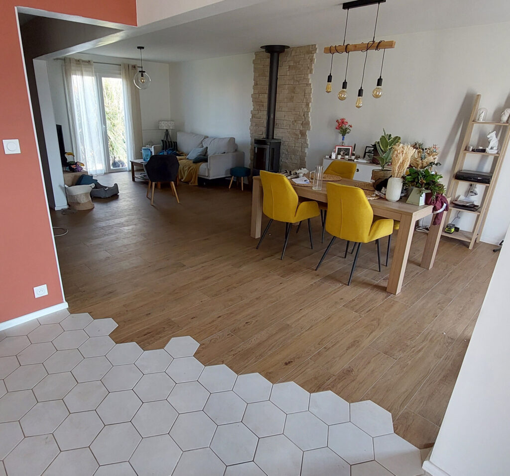 Salon et salle à manger décloisonnés avec un sol en bois et en carreaux hexagonaux fabriqués par un spécialiste du carrelage, des chaises jaunes autour d'une table en bois, un canapé gris et un poêle à bois adossé à un mur en pierre.