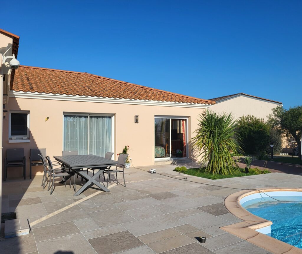 Maison de plain-pied avec un toit en tuiles, des portes coulissantes en verre, des meubles de jardin et une piscine lors d'une journée ensoleillée. Une terrasse carrelage grand format Vendée agrémente l'espace extérieur, bordé d'arbustes et d'une petite pelouse près de la piscine.