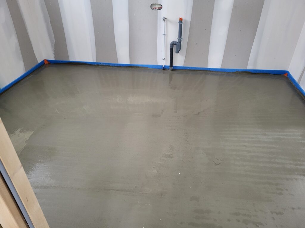 Sol en béton fraîchement coulé et nivelé dans une pièce avec des cloisons sèches non finies et des tuyaux de plomberie exposés sur le mur du fond.