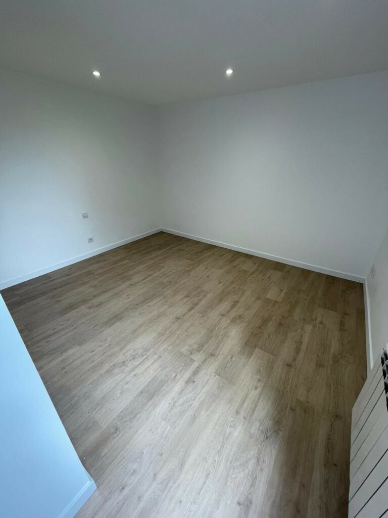 Pièce vide aux murs blancs, plafonniers encastrés, sol clipsé stratifié en bois clair et prises électriques apparentes sur les murs.
