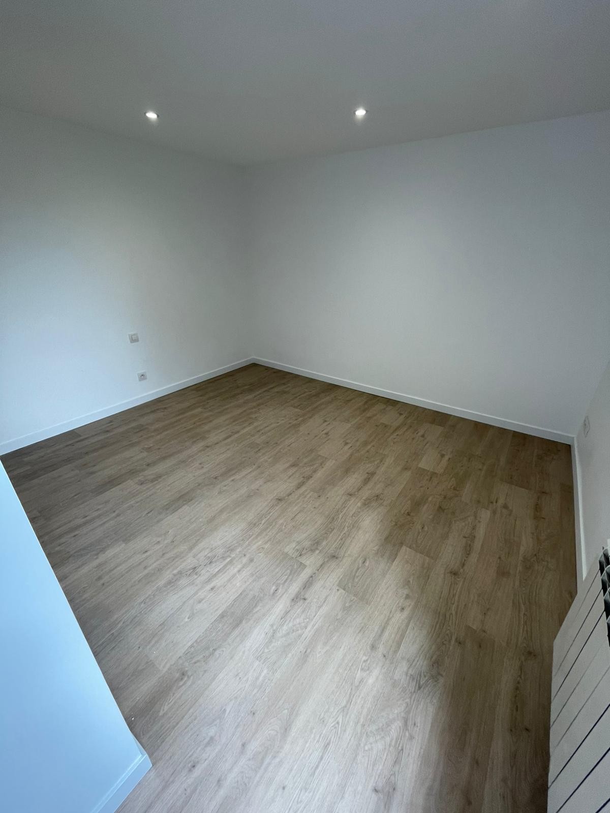 Pièce vide aux murs blancs, plafonniers encastrés, sol clipsé stratifié en bois clair et prises électriques apparentes sur les murs.