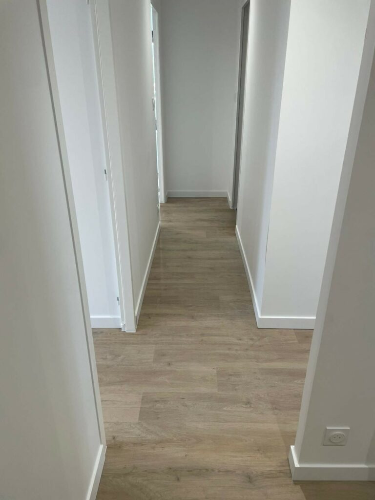 Un couloir étroit avec un sol clipsé en effet bois clair, des murs blancs et des portes blanches de chaque côté, menant à une zone plus lumineuse au bout.