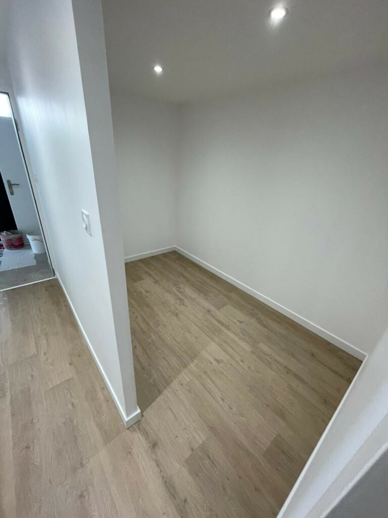 Une petite pièce vide aux murs blancs, au sol en stratifié clipsé effet bois, avec deux plafonniers et une porte ouverte menant à une autre zone.