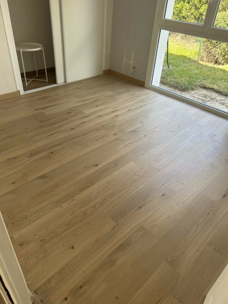 Chambre avec sol clipsé stratifié effet bois clair récemment installé, grande fenêtre avec vue sur l'extérieur, murs blancs unis et petite table ronde dans le coin.