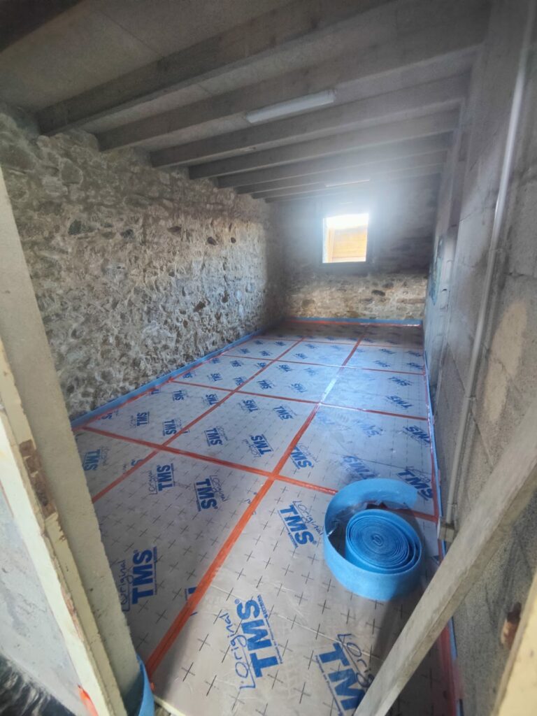 Une petite pièce avec des murs en pierre et une seule fenêtre, le sol recouvert de plaques d'isolation et un rouleau de mousse bleue dans un coin.
