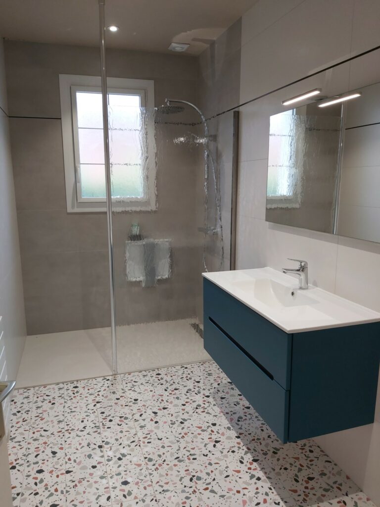 Salle de bains moderne avec sol en terrazzo, cabine de douche en verre, meuble-lavabo bleu fixé au mur, miroir rectangulaire et fenêtre apportant de la lumière naturelle.