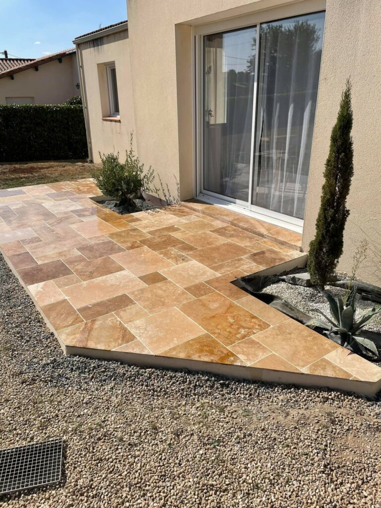 Une Terasse Travertin naturel beige et marron est adjacent à une maison avec des portes vitrées coulissantes, de petites plantes et des bordures en gravier.